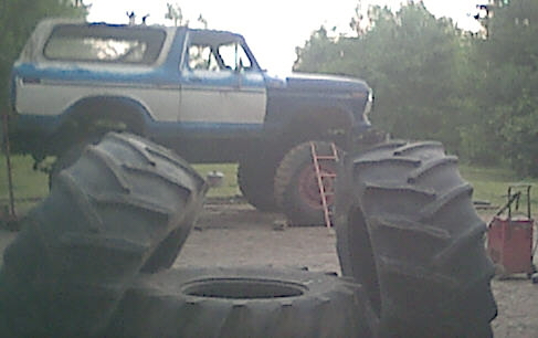 Blue and white Ford Bronco on huge tractor tires, side view, with large tires in foreground