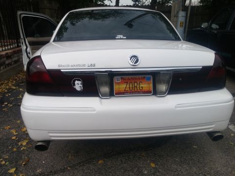 2000 Mercury Grand Marquis - The rear of a white 2000 Mercury Grand Marquis LSE features a custom license plate and a sticker of a man's face on the trunk.