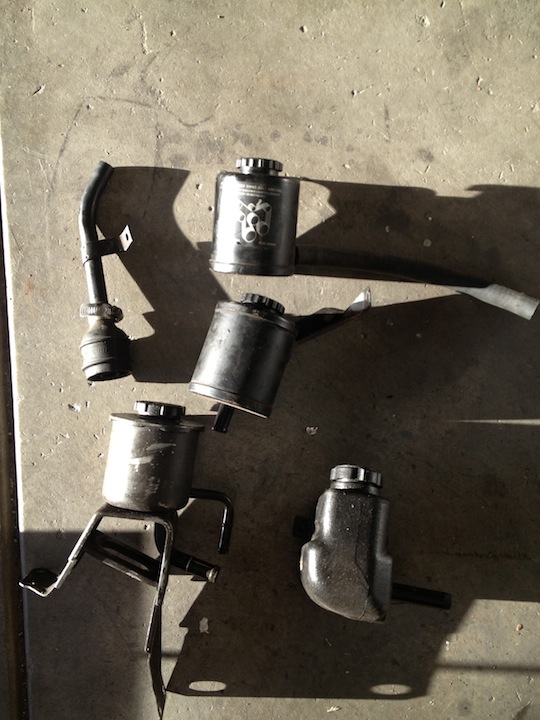 Four black automotive fluid reservoirs and a bent metal tube on a concrete surface
