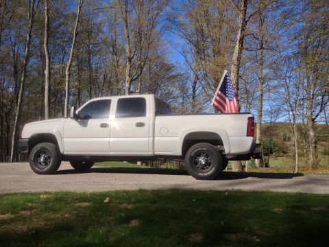 2006 Chevrolet Silverado 2500 - A white Chevrolet Silverado 2500 pickup truck is parked on a road with trees in the background and an American flag in the bed.
