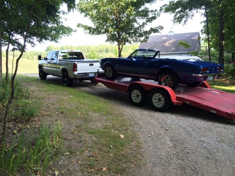 2006 Chevrolet Silverado 2500 - Silverado pickup tows a blue classic convertible on a trailer down a gravel driveway.