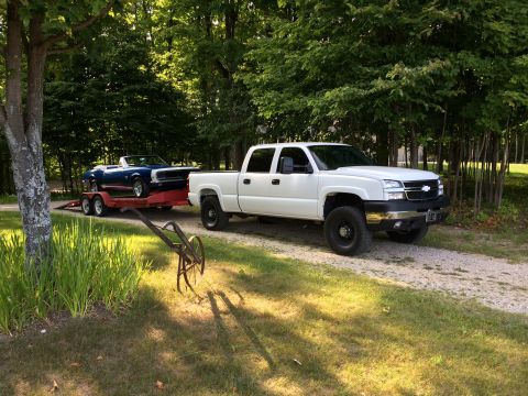 2006 Chevrolet Silverado 2500 - A white 2006 Chevrolet Silverado 2500 tows a blue convertible down a gravel driveway surrounded by trees.