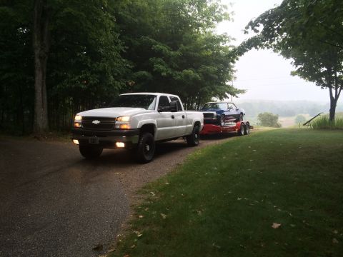 2006 Chevrolet Silverado 2500 - A white 2006 Chevrolet Silverado 2500 pickup truck is seen from a 3/4 front view, towing a blue classic car on a trailer.