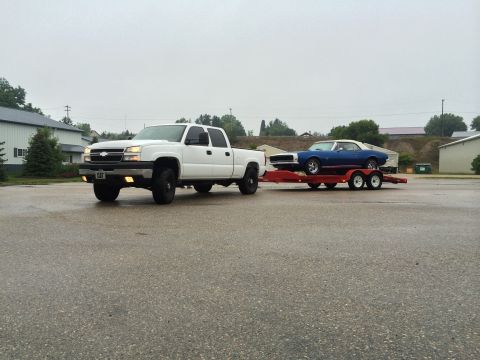 2006 Chevrolet Silverado 2500 - A white pickup truck tows a blue classic car on a trailer under an overcast sky.