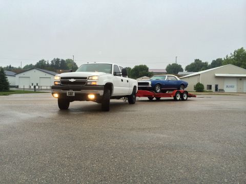2006 Chevrolet Silverado 2500 - A white Chevrolet pickup truck tows a blue classic car on a trailer in front of industrial buildings.