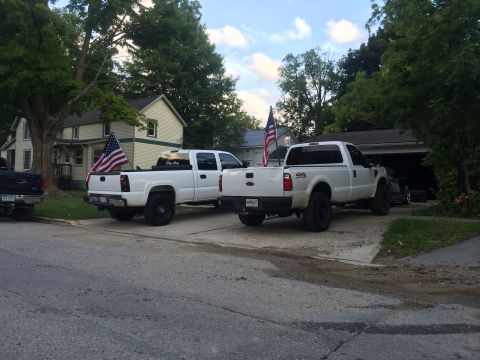 2006 Chevrolet Silverado 2500 - Two white pickup trucks are parked in a driveway, one displaying an American flag in its bed.