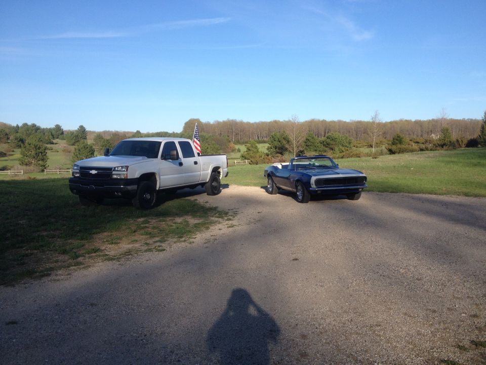 White pickup truck with American flag in bed parked next to a blue convertible on a gravel driveway