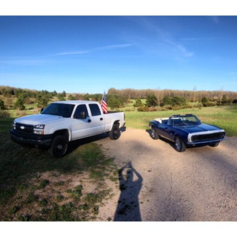 2006 Chevrolet Silverado 2500 - A white 2006 Chevrolet Silverado 2500 pickup truck and a blue convertible are parked on a dirt road with trees in the background.