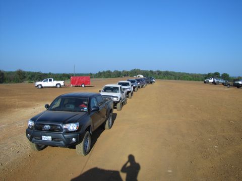 1995 Ford Bronco - Multiple trucks and SUVs are lined up on a dirt road in an open, dry field on a sunny day.
