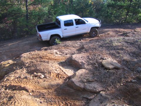 1995 Ford Bronco - White Toyota Tacoma pickup truck driving on a challenging off-road trail with rocky terrain and dirt mounds.