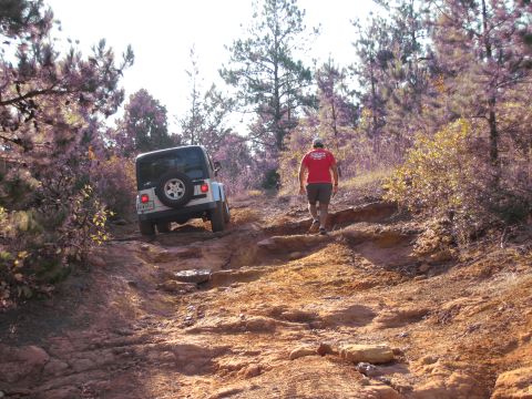 1995 Ford Bronco - A white Jeep Wrangler navigates a challenging rocky trail through a wooded area, with a person walking ahead on the path.