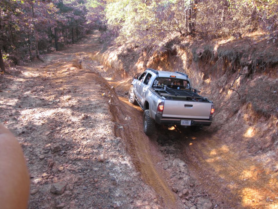 Silver pickup truck on a dirt trail, rear view. The truck has a rack in the bed and is driving off-road