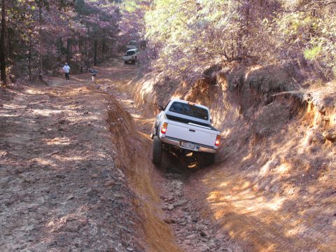 1995 Ford Bronco - Off-roading scene with a white pickup truck navigating a rutted dirt path, with another vehicle in the distance.