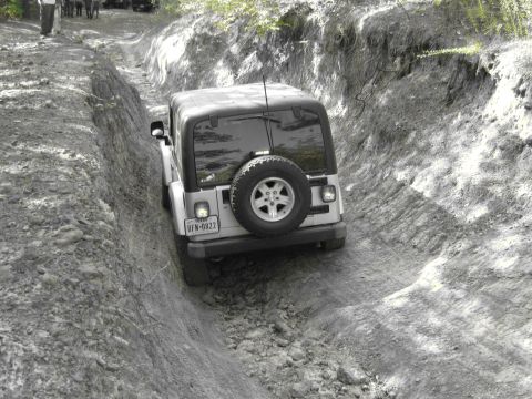 1995 Ford Bronco - A Jeep Wrangler TJ navigates a narrow, rutted dirt path, showing its rear and spare tire.