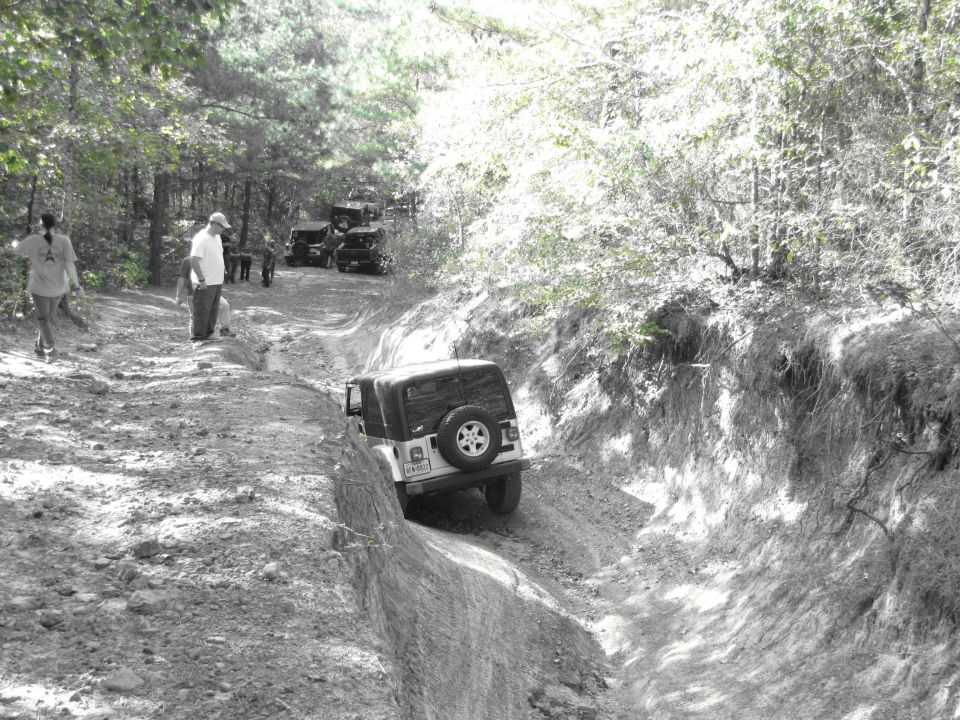 Several Jeeps navigate a steep, muddy off-road trail with trees on either side. Two people stand watching on the left