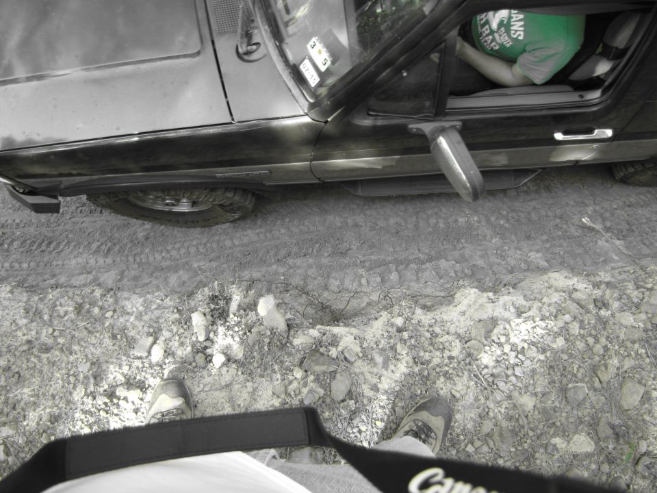 Black Ford Bronco side view, driver's door open, person in green shirt visible inside. Muddy ground with tire tracks