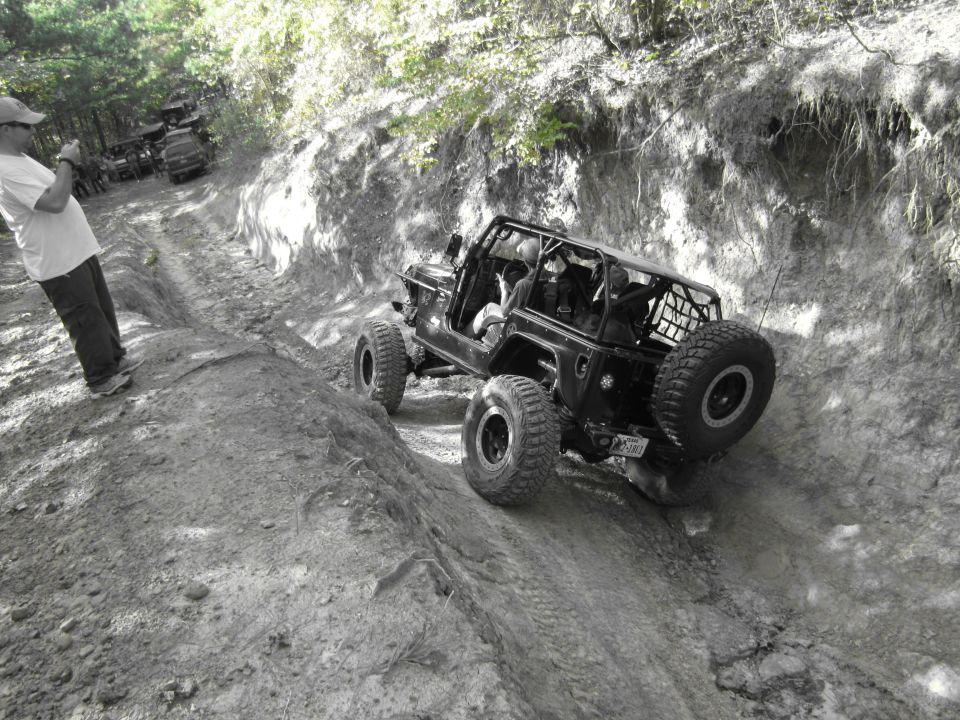 Black Jeep Wrangler with a roll cage and spare tire on a steep dirt trail, with a person taking a photo on the left