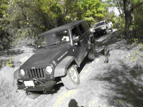 1995 Ford Bronco - Two off-road vehicles navigate a challenging dirt trail with dense foliage on either side.