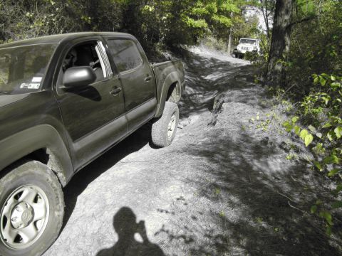 1995 Ford Bronco - Two off-road vehicles navigate a narrow dirt path surrounded by dense foliage.