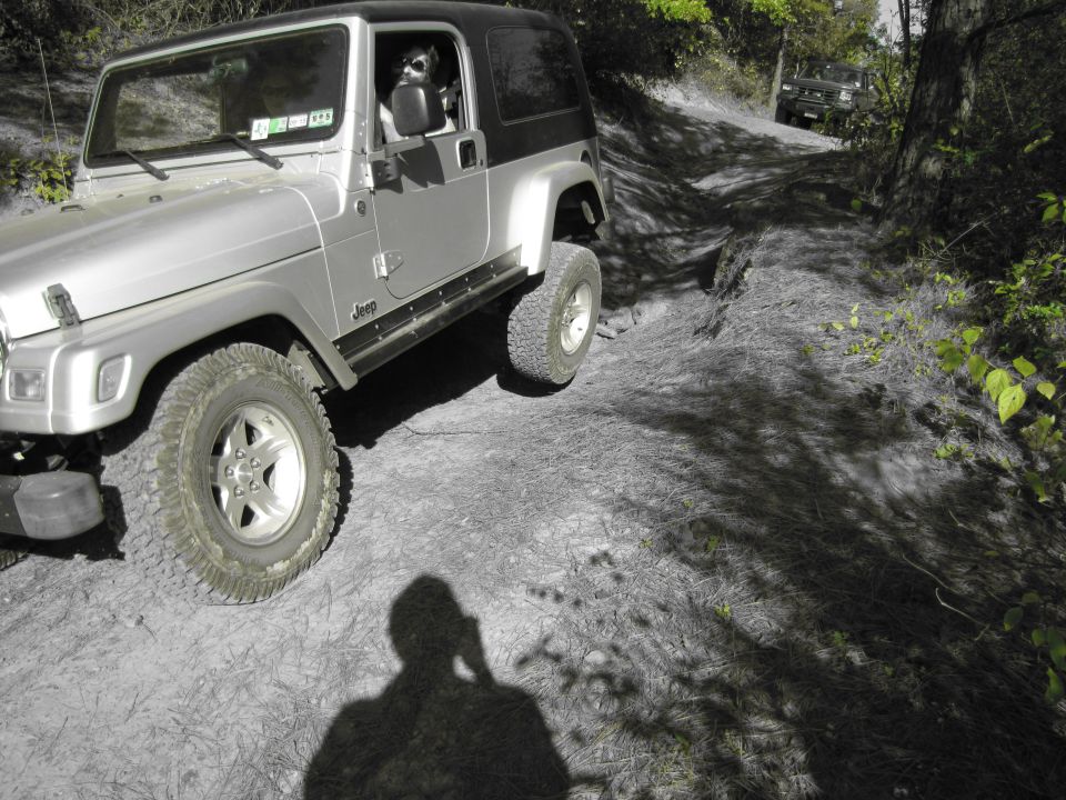 Silver Jeep Wrangler with black top on a dirt trail, driver visible, another vehicle in background
