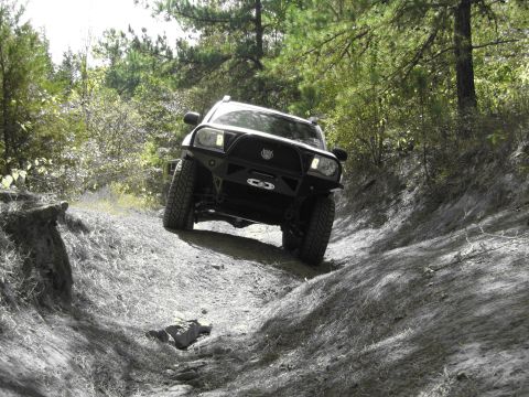 1995 Ford Bronco - An off-road SUV navigates a steep, rutted dirt path surrounded by trees.