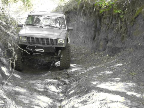 1995 Ford Bronco - A Jeep Cherokee XJ navigates a narrow, muddy trail with steep dirt banks.