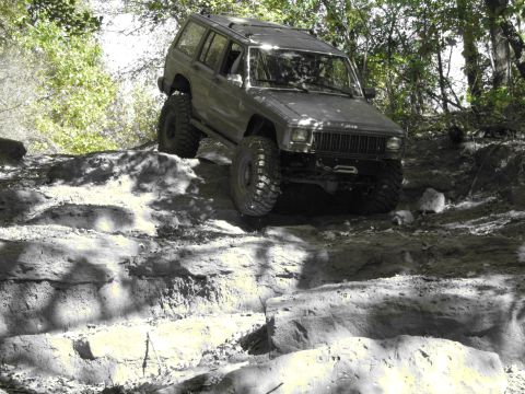 1995 Ford Bronco - A gray Jeep Cherokee XJ navigates a challenging rocky trail with its front-left tire lifted high.