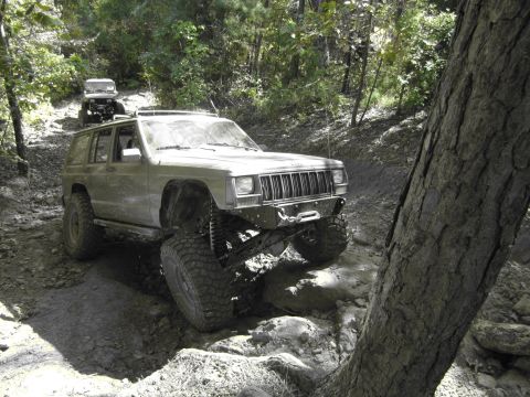 1995 Ford Bronco - Two modified Jeeps tackle a challenging off-road path in a wooded area.