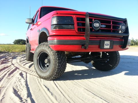 1995 Ford Bronco - This red Ford Bronco, modified for off-road use, sits on a sandy surface under a clear blue sky.