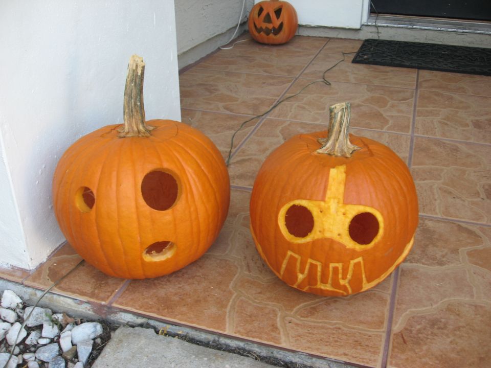 Two carved pumpkins on a tiled porch, one with a surprised face, the other with a robot-like face