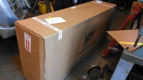1989 Ford Bronco - A large cardboard box labeled LMC Truck sits on a workshop floor next to other automotive parts.