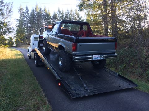 1989 Ford Bronco - A tow truck is hauling a lifted Ford Bronco on a rural road.