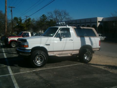 1996 Ford Bronco - A white 1996 Ford Bronco with a tan top and roof-mounted lights is parked in a lot, with another truck nearby.