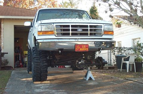 1995 Ford Bronco - Front view of a white lifted Ford truck on jack stands in a driveway with the passenger-side front wheel and hub removed, exposing the soli…