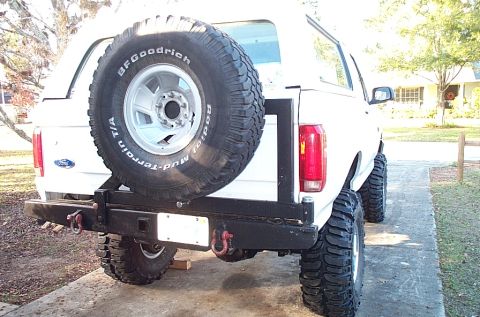 1995 Ford Bronco - Three-quarter rear view of a white lifted Ford Bronco parked on a concrete driveway, with a black tube rear bumper, D-ring shackles, and a…