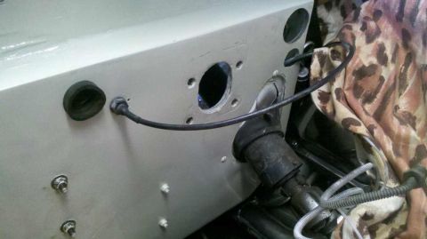 1982 Ford Bronco - A close-up view of the firewall of a vehicle, showing a large circular opening and a rubber grommet with a cable passing through it. Severa…