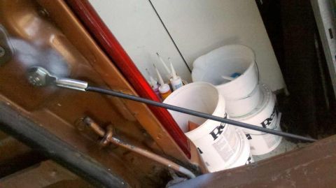 1982 Ford Bronco - A close-up view of a metal cable attached to the interior of a vehicle door, with several buckets and bottles of sealant in the background.