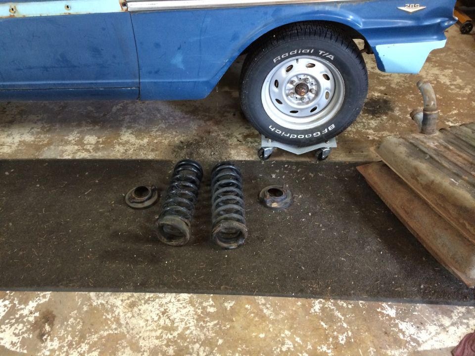 Two front coil springs and upper mounts on a mat next to a blue vintage car and a rusty fuel tank.