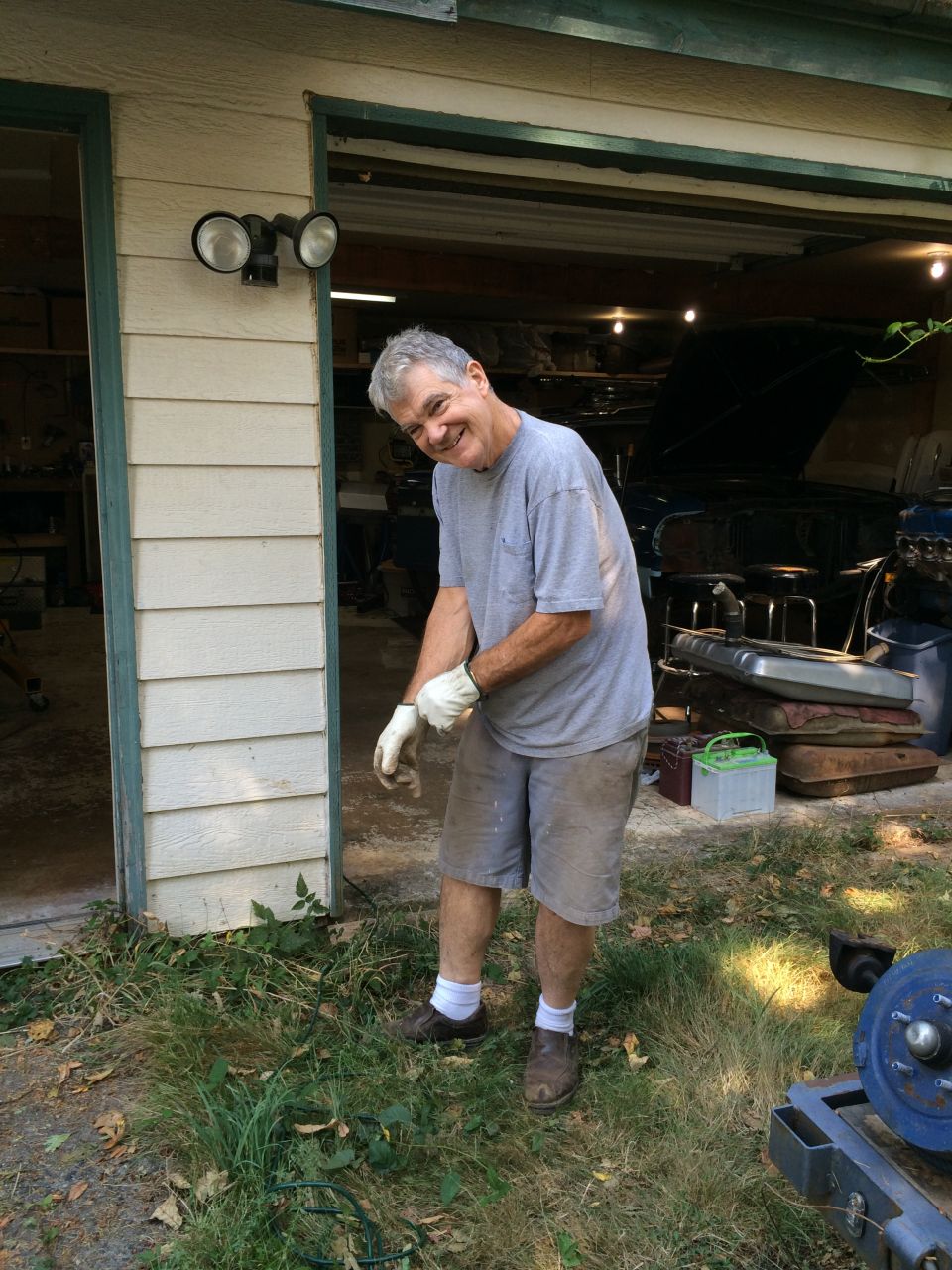 Man in grey t-shirt and shorts smiling in front of an open garage with a car inside.