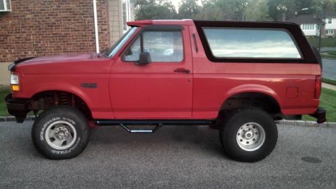 1994 Ford Bronco - Side profile of a red Ford Bronco parked on a paved driveway. The vehicle features aftermarket side steps with LED lights and large off-roa…