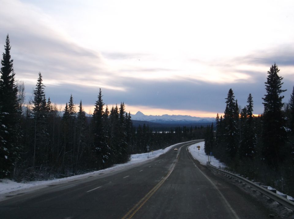 A road through a snowy forest with mountains in the distance.