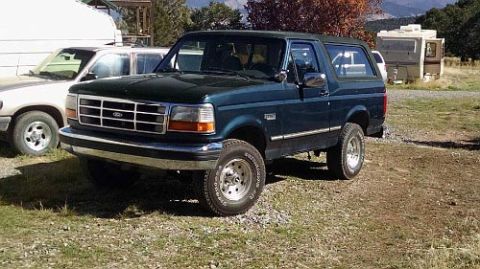 1994 Ford Bronco - A dark green 1994 Ford Bronco is parked on a gravelly lot next to a white trailer. The front of the SUV is facing the camera at a three-qua…