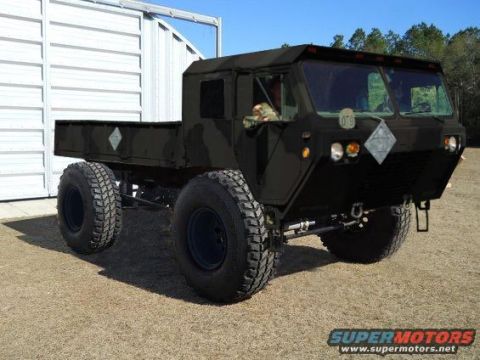 1994 Ford Bronco - black military-style heavy tactical truck with a HEMTT-type cab on large off-road tires parked beside a metal building.