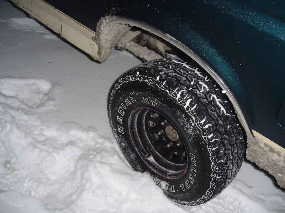 Snow-packed all-terrain tire on black steel wheel, green truck at night