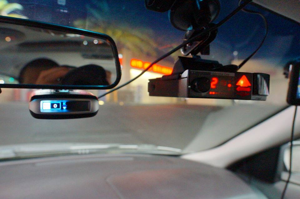 Radar detector with red lights and a dashcam mounted on a windshield, with a rearview mirror reflecting a person