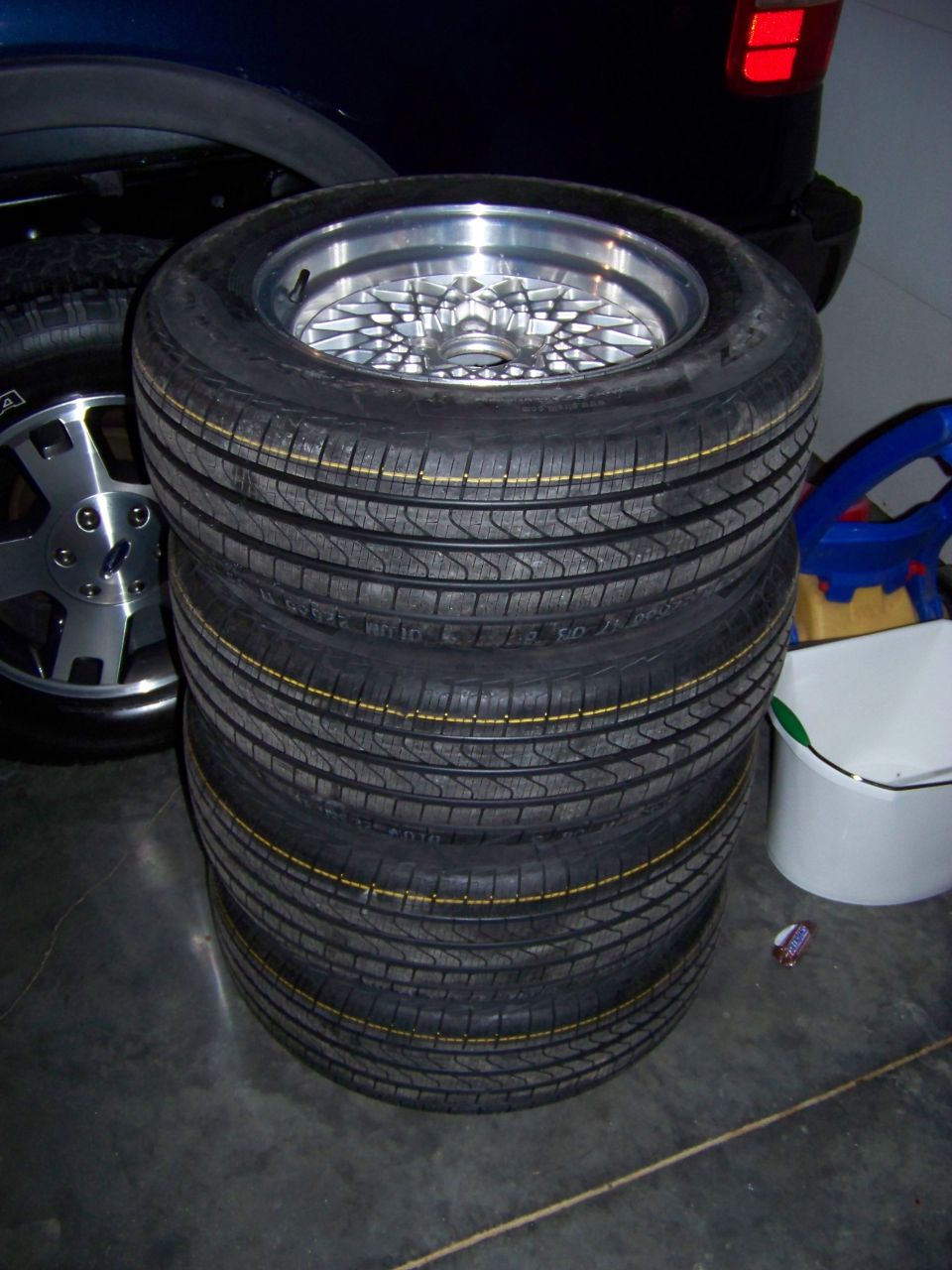 Stack of four tires with a silver mesh wheel on top, next to a dark blue vehicle's rear quarter panel