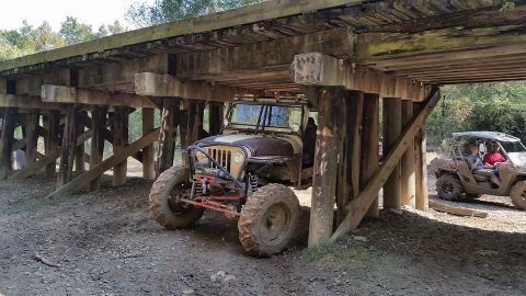 Modified Jeep parked under a wooden bridge, another UTV visible to the right