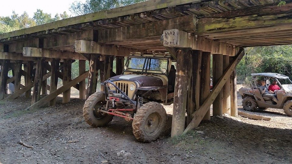 Modified Jeep parked under a wooden bridge, another UTV visible to the right