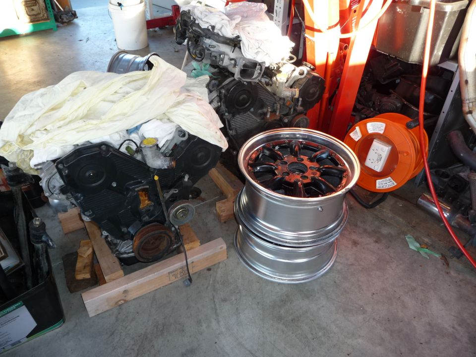 Two car engines, one partially covered, sit on wooden blocks next to a stack of three shiny aftermarket wheels