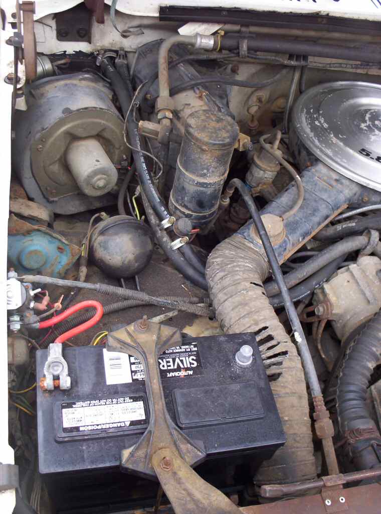 Grimy engine bay corner with battery, alternator and 5.8 air cleaner edge under white hood